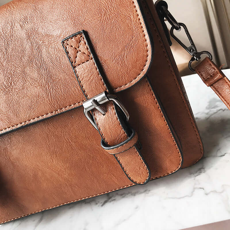 Close-up of a stylish commuter messenger bag in brown PU leather with buckle detailing on a marble surface.